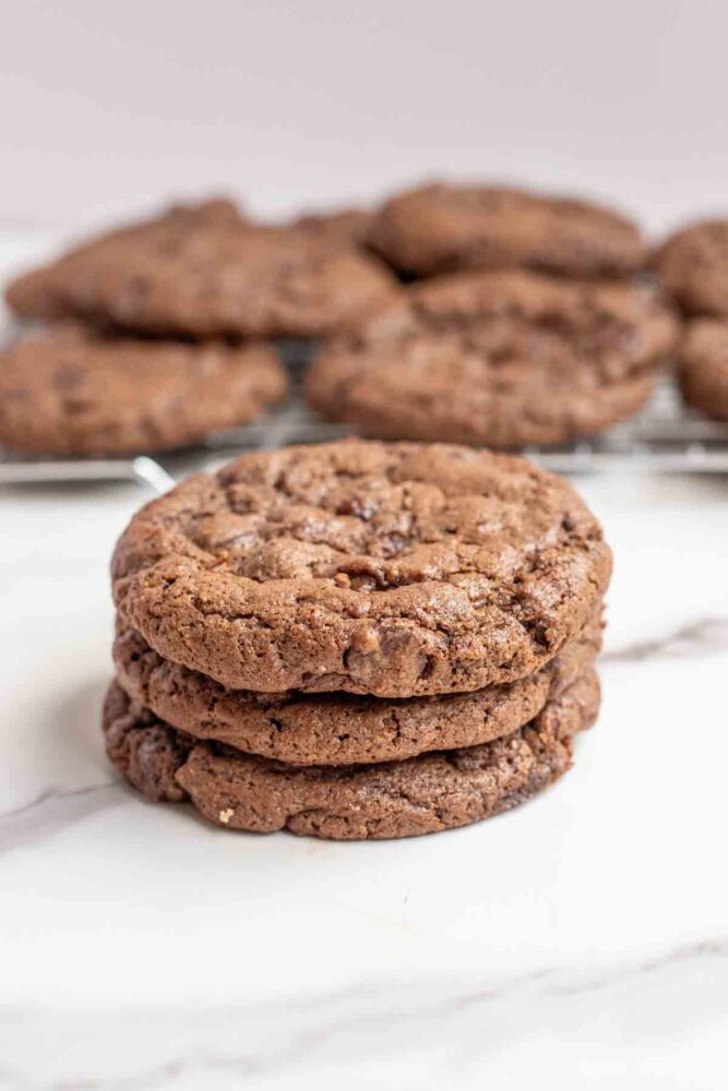 Three sourdough chocolate cookies stacked on top of each other on a marble countertop with more cookies in the background.
