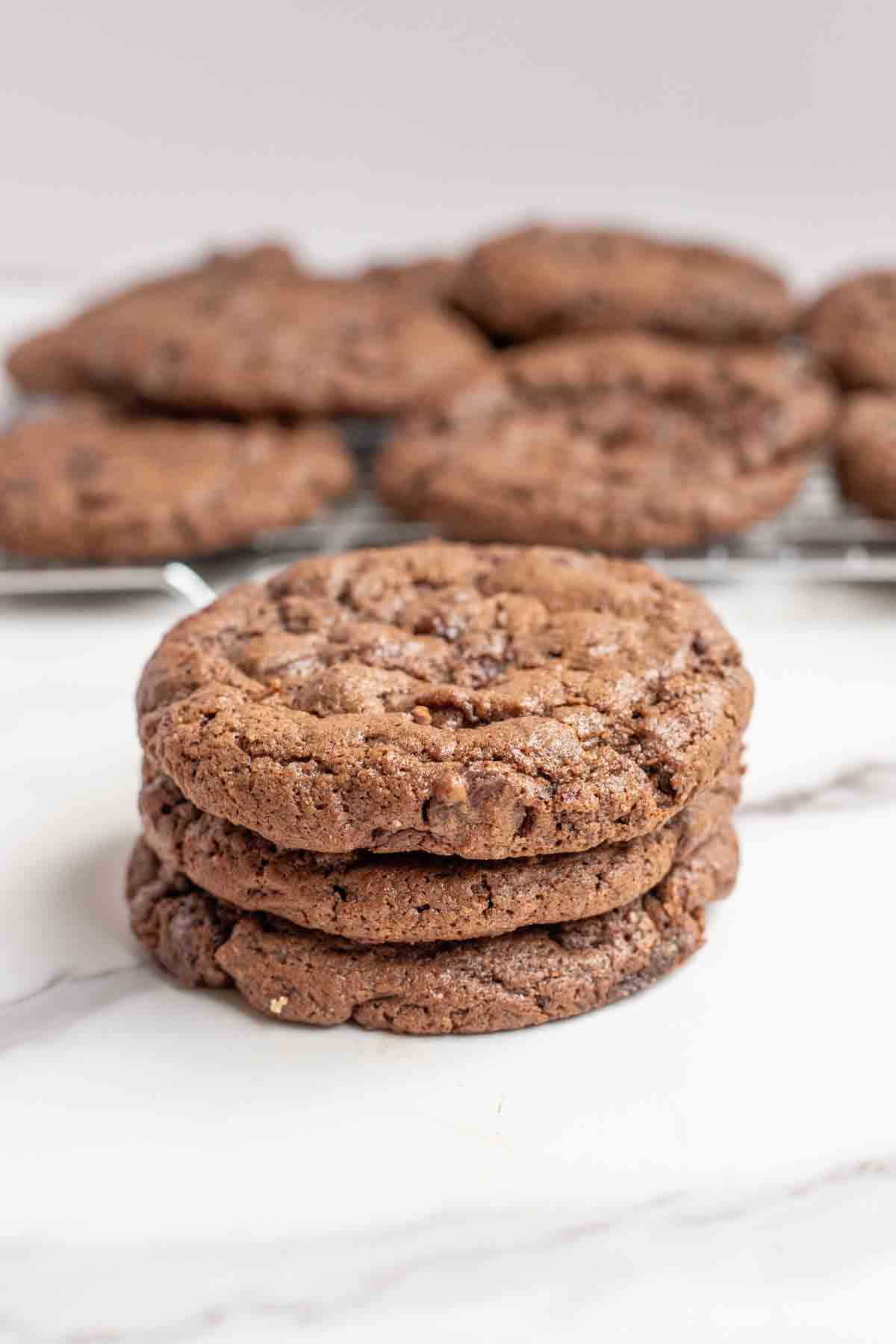 Three sourdough chocolate cookies stacked on top of each other on a marble countertop with more cookies in the background.