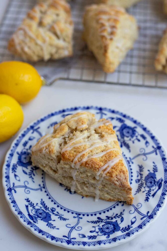 sourdough lemon scone drizzled with lemon icing on a vintage blue and white plate with lemons and scones in the background.