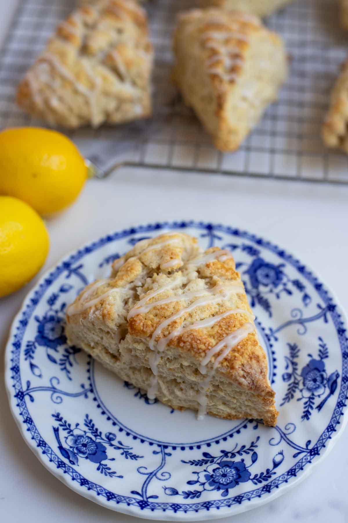 sourdough lemon scone drizzled with lemon icing on a vintage blue and white plate with lemons and scones in the background.