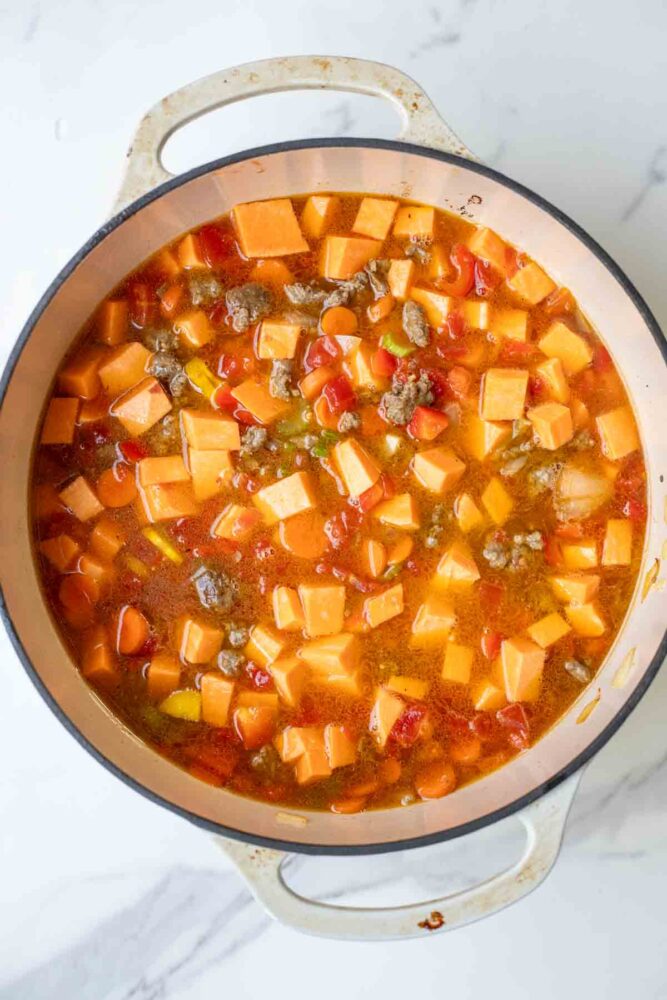 Broth, sweet potatoes, and tomatoes added to a pot of meat and vegetables.