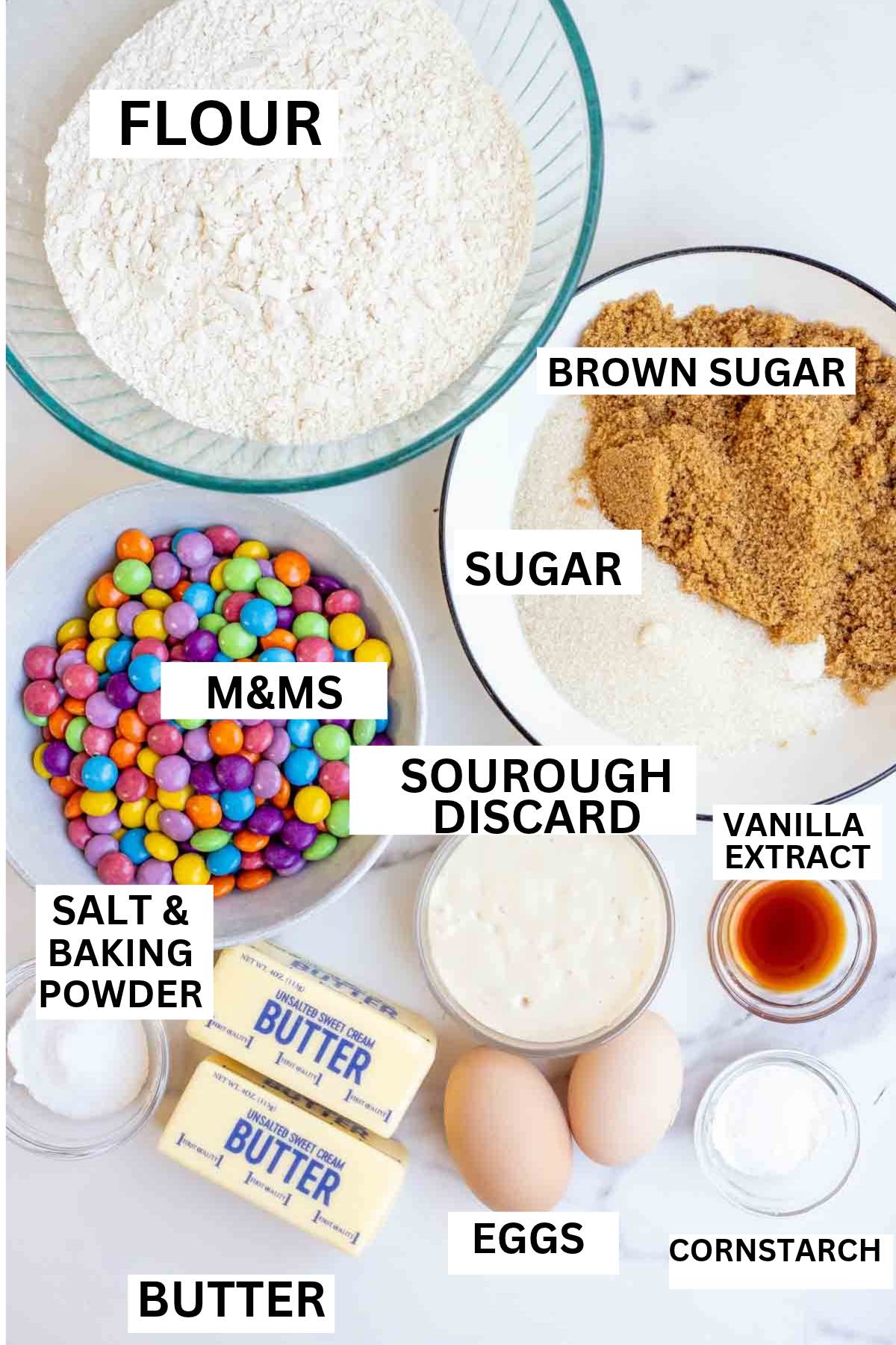 Flour, butter, m&ms, sourdough starter and more on a marble countertop.