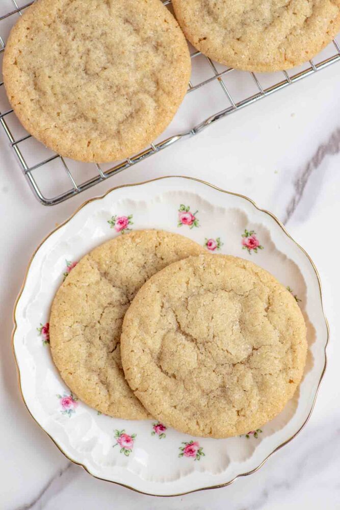 Two sugar cookies on a floral plate with more cookies on a cooling rack in the background.